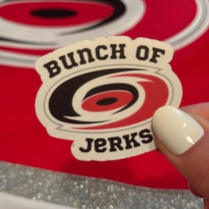 May include: A sticker with the text "Bunch of Jerks" in black letters. The sticker has a white border and features a red, black, and white logo. The background is red with a blurred logo.