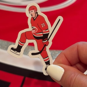 May include: A white sticker of a hockey player wearing a red and black jersey with the number 20 on it. The player is holding a hockey stick and is in a skating position.