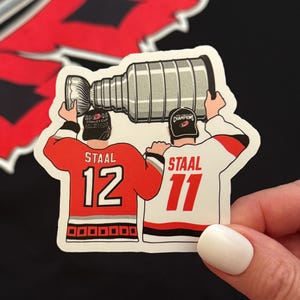 May include: A die-cut sticker featuring two hockey players in jerseys with the name STAAL and numbers 12 and 11, both wearing black caps and lifting a silver Stanley Cup trophy above their heads. The design includes red, white, and black colors.