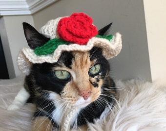 May include: A calico cat wearing a white crocheted sun hat with a red crocheted rose on top. The cat is lying on a white fluffy surface.