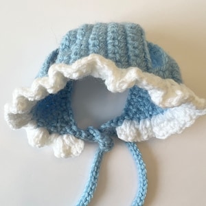 May include: A light blue crocheted baby bonnet with a white crocheted ruffle around the brim and a long tie.