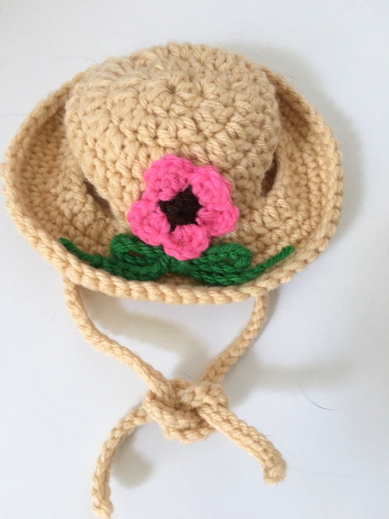 May include: A crocheted hat in a light beige color with a pink flower and green leaves. The hat has a long tie that can be tied under the chin.