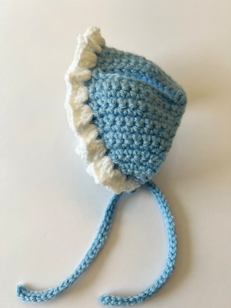 May include: A light blue crocheted baby hat with a white crocheted ruffle around the brim and a long blue crocheted tie.