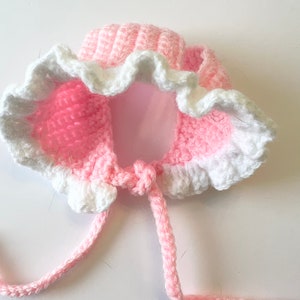 May include: Pink and white crocheted hat with a ruffled edge and ties. The hat is designed for a small child.