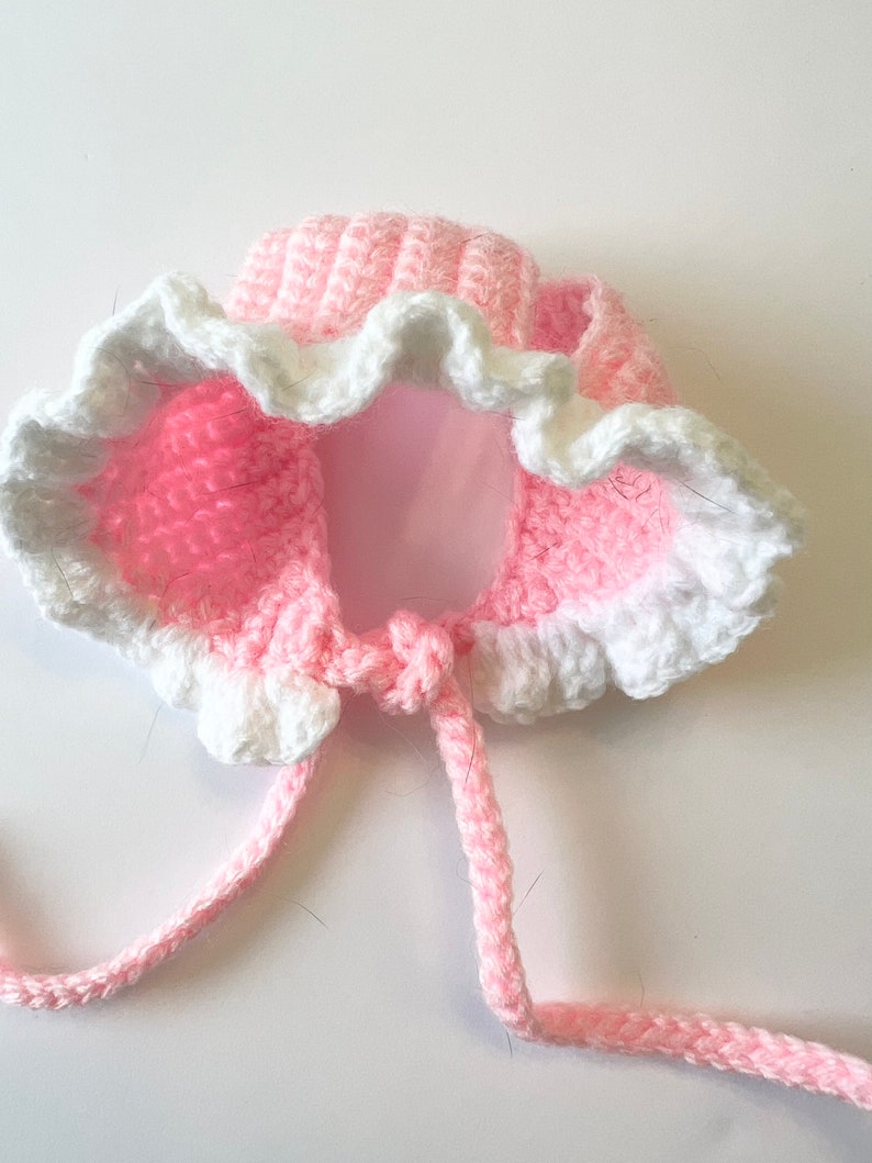 May include: Pink and white crocheted hat with a ruffled edge and ties. The hat is designed for a small child.