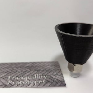 Pode incluir: Um objeto preto em forma de cone com um encaixe de metal. O objeto é provavelmente um protótipo, como indicado pelo cartão de visita com o texto "Tranquillity Protypes". O objeto está sobre um fundo branco.