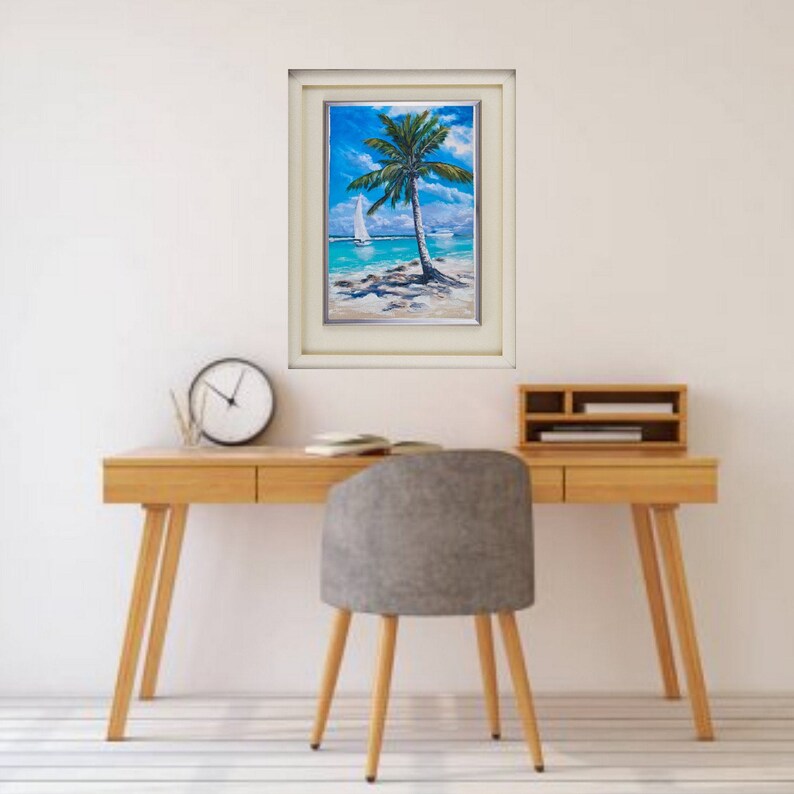 Puede incluir: Una pintura enmarcada de una escena tropical con una palmera en una playa con un velero en la distancia. La pintura est&aacute; en un marco blanco con un passe-partout crema.