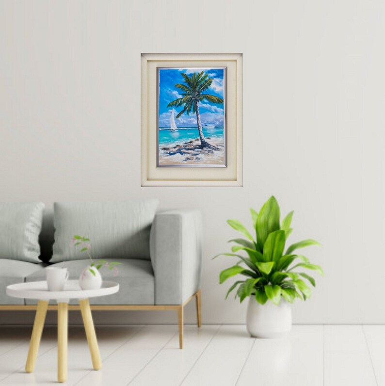 Puede incluir: Una pintura enmarcada de una palmera en una playa con un velero en la distancia. La pintura est&aacute; en un marco blanco con un borde plateado.