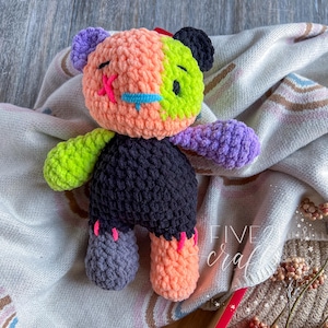 May include: A handmade crochet bear with a patchwork design. The bear has a black body, with a head split into orange, green, and purple sections. It has pink and blue facial details and grey and orange feet.