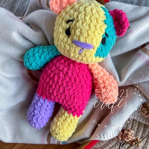 May include: A handmade, colourful crochet bear with a yellow head, pink body, and various coloured limbs. The bear has black eyes and a purple nose. The text "FIVE 21 crafts" is visible on the right side of the image.