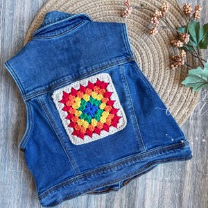 May include: A sleeveless denim vest with a colorful, crocheted square patch. The patch features a rainbow of colors including red, orange, yellow, green, blue, and purple. The vest is a medium wash denim and has a classic collar.