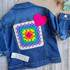 May include: A blue denim jacket with a large, colorful crochet square and a pink heart attached to the back. The crochet square features a rainbow of colors. The jacket has a Soho Blue Denim tag.
