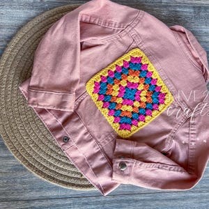 May include: A dusty rose denim jacket with a colorful crochet patch. The patch is square, featuring a vibrant pattern of pink, blue, yellow, and orange. The jacket is buttoned and lies on a woven placemat. The text "LIVE 21 crafts" is visible.