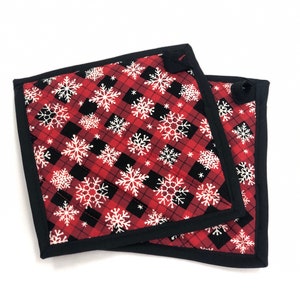 May include: Two red and black plaid pot holders with white snowflake designs and black trim.