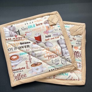May include: Two beige quilted pot holders with a coffee-themed print. The print includes coffee-related words and illustrations of coffee cups, a French press, and coffee beans. The edges are trimmed with a solid beige fabric.
