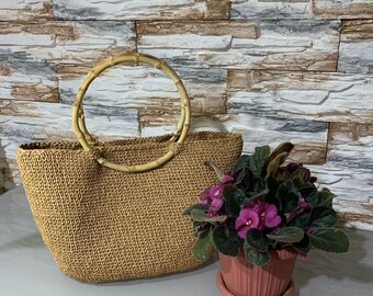 May include: A brown woven tote bag with a natural bamboo handle. The bag is sitting on a textured surface with a small potted plant with purple flowers.
