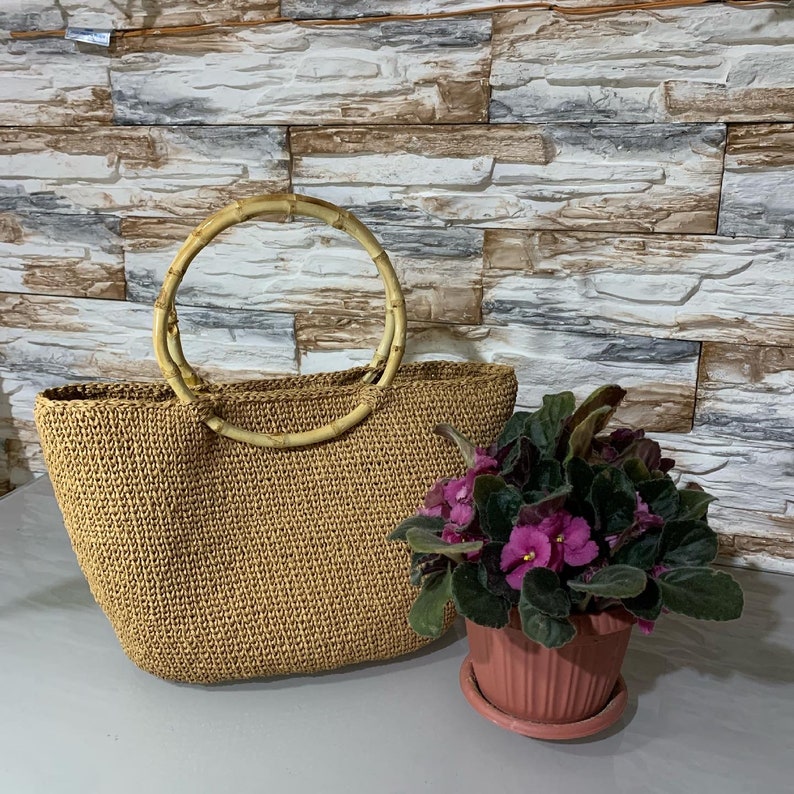 May include: A brown woven tote bag with a natural bamboo handle. The bag is sitting on a textured surface with a small potted plant with purple flowers.