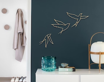 Op de afbeelding: Drie gouden metalen vogelwanddecoraties in minimalistische stijl, gemonteerd op een donkerblauwe muur. De vogels zijn in vlucht, met hun vleugels uitgespreid. De decoraties zijn een moderne en stijlvolle manier om een vleugje natuur aan elke kamer toe te voegen.