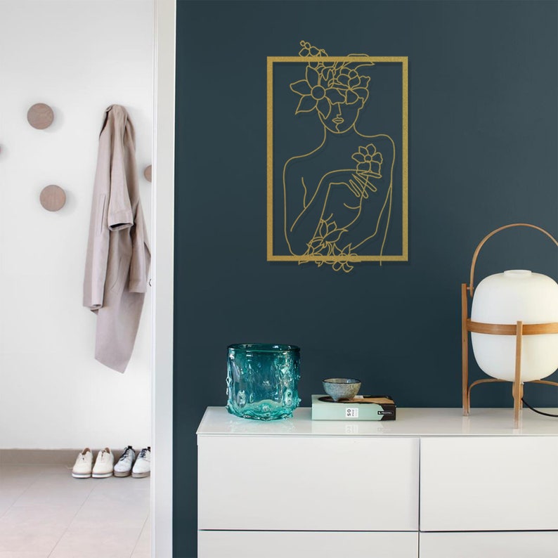 Pu&ograve; includere: Un disegno a contorno dorato di una donna con fiori nei capelli e sul petto, incorniciato in un rettangolo dorato, su una parete blu scuro.