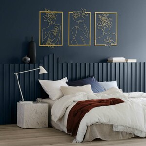 Pu&ograve; includere: Tre sculture da parete in metallo dorato con un design a linea d'arte di un viso femminile con fiori tra i capelli. Le sculture sono appese a una parete blu scuro sopra un letto bianco con una coperta marrone.