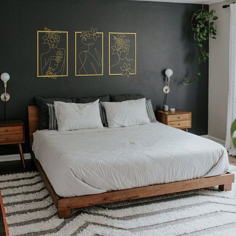 Pu&ograve; includere: Tre pezzi d'arte da parete in metallo dorato con disegni lineari di donne con copricapi floreali. L'arte &egrave; esposta su una parete grigio scuro sopra una struttura letto in legno con biancheria da letto bianca.