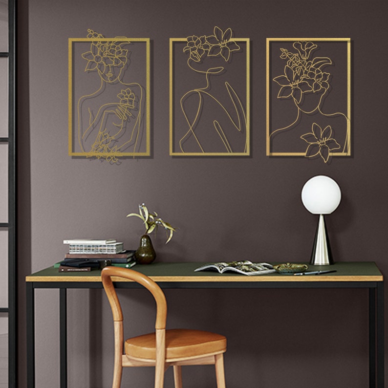 Pu&ograve; includere: Tre pezzi di arte da parete in metallo dorato con un disegno a linea di un viso di donna con fiori nei capelli. I pezzi d'arte sono appesi a una parete grigio scuro.
