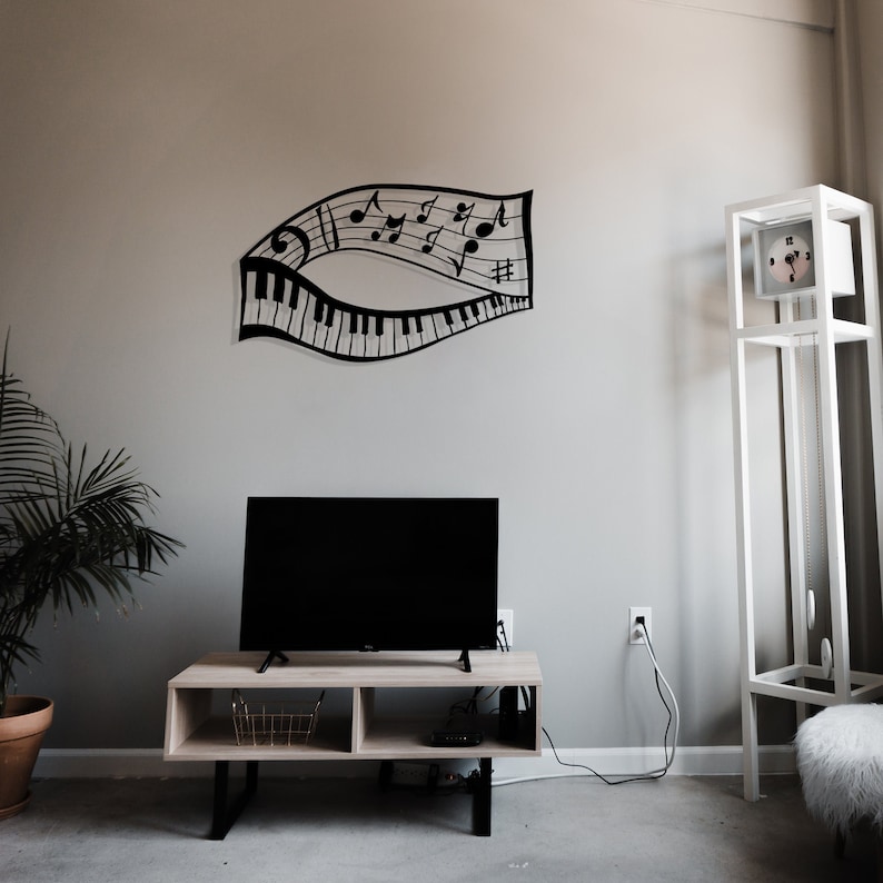 May include: A black metal wall decor featuring musical notes and a piano keyboard. The decor is mounted on a light gray wall above a light wood entertainment center with a black flat screen TV. A white clock tower stands in the corner of the room.