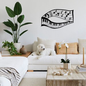 May include: Black metal wall art featuring a piano keyboard and musical notes in a flowing design. The art is displayed on a white wall above a white couch with a white dog and plants.
