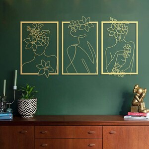 Pu&ograve; includere: Tre pezzi di arte da parete in metallo dorato con un disegno a linea di una persona con fiori nei capelli. Lo sfondo &egrave; una parete verde scuro.