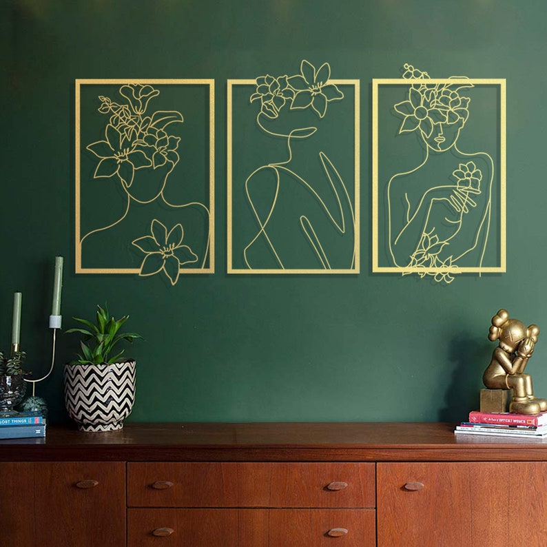 Pu&ograve; includere: Tre pezzi di arte da parete in metallo dorato con un disegno a linea di una persona con fiori nei capelli. Lo sfondo &egrave; una parete verde scuro.