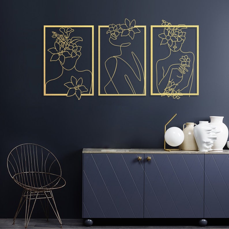 Pu&ograve; includere: Tre pezzi di arte da parete in metallo dorato con disegni lineari di donne con fiori nei capelli. L'arte &egrave; esposta su una parete blu scuro sopra un armadio blu scuro con maniglie dorate.