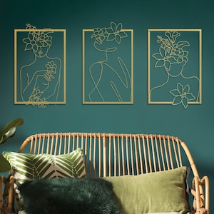 Pu&ograve; includere: Tre sculture da parete in metallo dorato che raffigurano una figura femminile stilizzata con fiori tra i capelli. Le sculture sono incorniciate in metallo dorato e sono posizionate su una parete verde turchese.