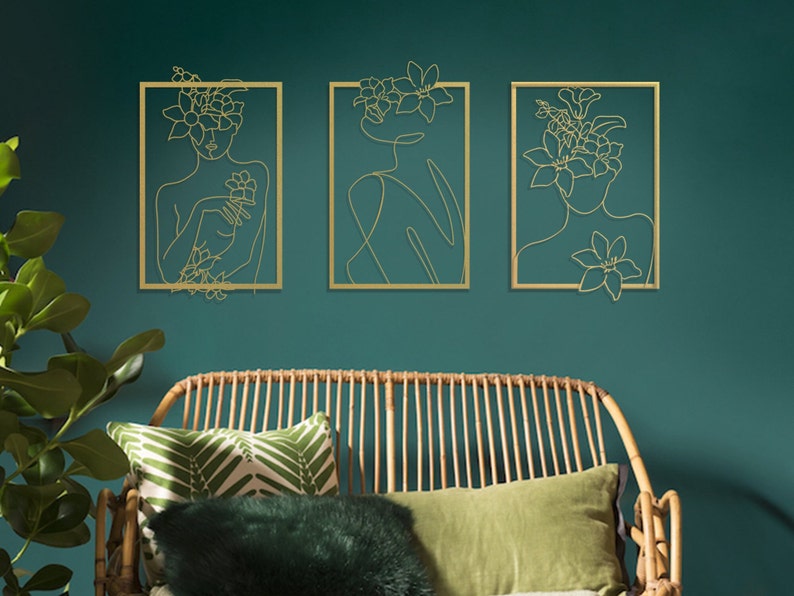 Pu&ograve; includere: Tre sculture da parete in metallo dorato che raffigurano una figura femminile stilizzata con fiori tra i capelli. Le sculture sono incorniciate in metallo dorato e sono posizionate su una parete verde turchese.