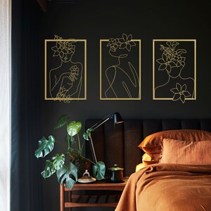 Pu&ograve; includere: Tre stampe di arte lineare dorate incorniciate di donne con accenti floreali su una parete nera. Le stampe sono in un ambiente da camera da letto con un letto marrone e un comodino marrone.