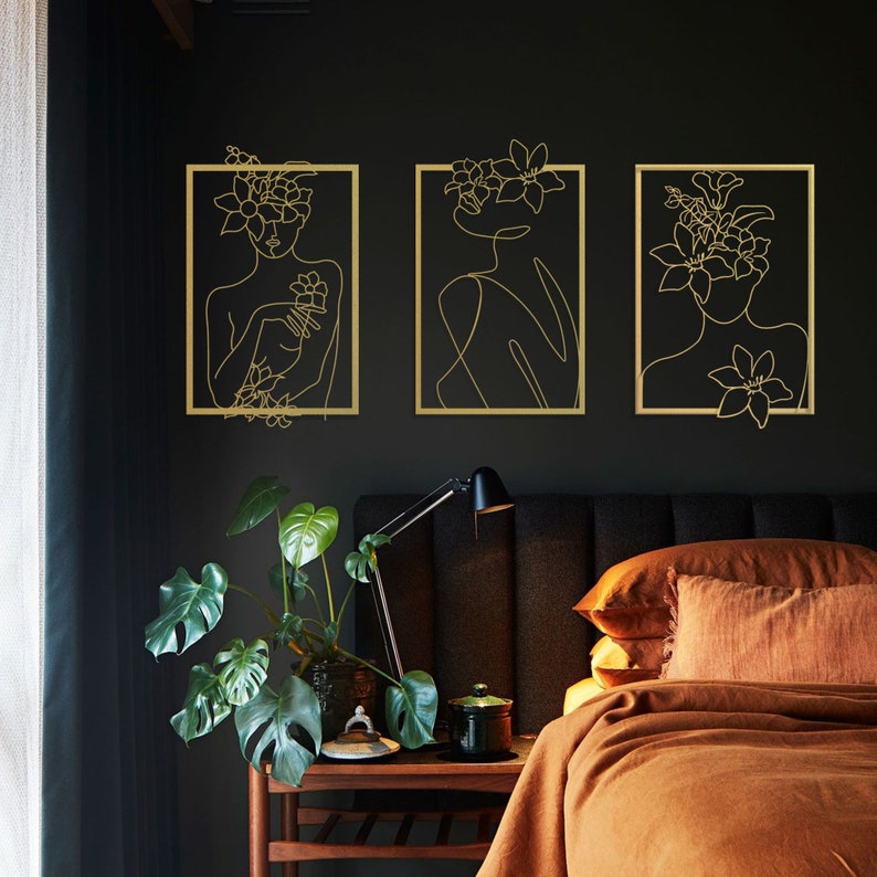 Pu&ograve; includere: Tre stampe di arte lineare dorate incorniciate di donne con accenti floreali su una parete nera. Le stampe sono in un ambiente da camera da letto con un letto marrone e un comodino marrone.