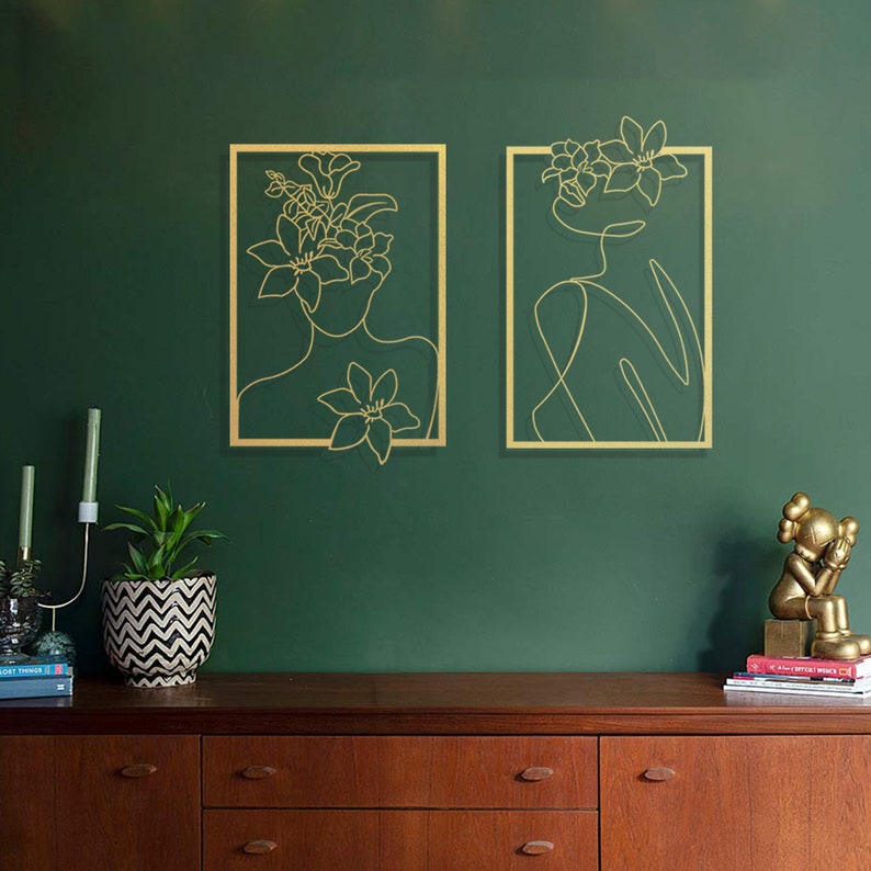 Pu&ograve; includere: Due pezzi di arte da parete in metallo dorato con un disegno a linea di una persona con fiori nei capelli. L'arte &egrave; appesa a una parete verde scuro sopra una cassettiera in legno.