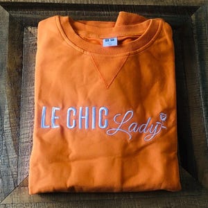 Puede incluir: Una sudadera naranja con la inscripción bordada "LE CHIC Lady" en escritura blanca. La sudadera tiene cuello redondo y una etiqueta en el cuello. La prenda está doblada cuidadosamente sobre una superficie de madera.