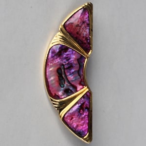 以下が含まれることがあります： 曲線的な金色のペンダント。紫とピンクの虹色のあわびの貝殻が象嵌されています。ペンダントは幾何学的なデザインで、3つの貝殻部分があり、金色の金属で縁取られ、細い線でアクセントがつけられています。