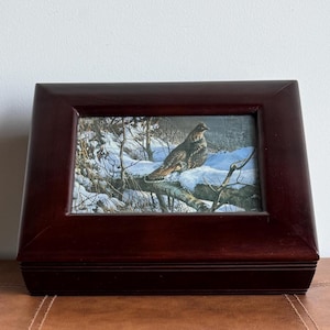 Puede incluir: Una caja rectangular de madera marrón oscuro con una imagen enmarcada de un pájaro posado en una rama cubierta de nieve. La obra de arte representa una escena invernal con árboles y nieve. La caja está sobre una superficie de cuero marrón.