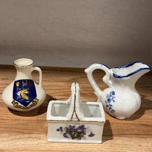 May include: Three ceramic decorative pieces: a small pitcher with a blue and white floral design, a small white vase with a crest, and a rectangular container with a handle and a floral design.