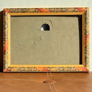May include: Rectangular picture frame with a decorative border featuring a pattern of brown, orange, and red shapes. The frame has a gold-coloured inner edge and a cardboard backing with a hanging tab.