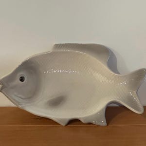 Puede incluir: Una fuente de cerámica con forma de pez en tonos grises y blancos. La fuente tiene una superficie texturizada, una cola detallada y un ojo oscuro. La fuente está sobre una superficie de madera.