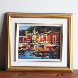 May include: Framed art print depicting a coastal Italian town with colorful buildings and boats in the water. The artwork is framed in a gold-colored wooden frame with a white matting. The painting captures the reflections in the water.
