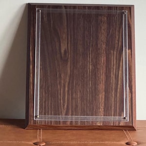 May include: A rectangular wooden plaque with a dark brown finish and a clear acrylic insert. The plaque is framed with a darker wood border and secured with small brass-colored pins. The wood grain is visible.