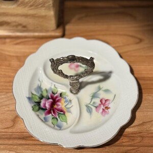 May include: A white, three-sectioned serving dish with a silver handle. The dish features a scalloped edge and is decorated with pink floral designs and green leaves. The handle is ornate and adds a vintage touch to the piece.