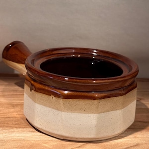 Puede incluir: Un cuenco de sopa de cerámica con asa, que presenta un diseño tricolor. El cuenco tiene un interior y borde marrón oscuro, una banda beige y una base color crema. El asa también es marrón. El cuenco está sobre una superficie de madera.