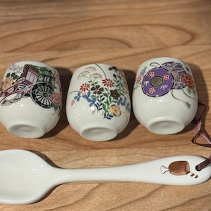 Może przedstawiać: Trzy małe, białe ceramiczne kubki z kolorowymi wzorami kwiatowymi i wózkami oraz pasująca łyżka. Łyżka ma brązowo-biały wzór w pobliżu uchwytu. Kubki i łyżka znajdują się na drewnianej powierzchni.