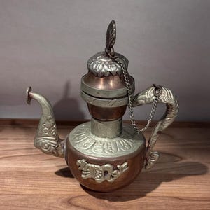 May include: An ornate, antique metal teapot with a copper-colored body and silver-colored accents. The teapot features a decorative spout, handle, and lid, with a chain connecting the lid to the handle. The design includes embossed patterns and a feather-shaped finial.
