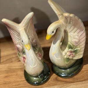 May include: A pair of ceramic swan ornaments. The swans are white with pink and beige detailing on their wings and bodies. They have yellow beaks and are decorated with pink flowers. The bases are green.