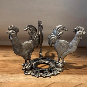 May include: A silver-coloured metal egg cup holder with two rooster figures. The holder has a circular base with floral detailing and a central support. The item is likely vintage.